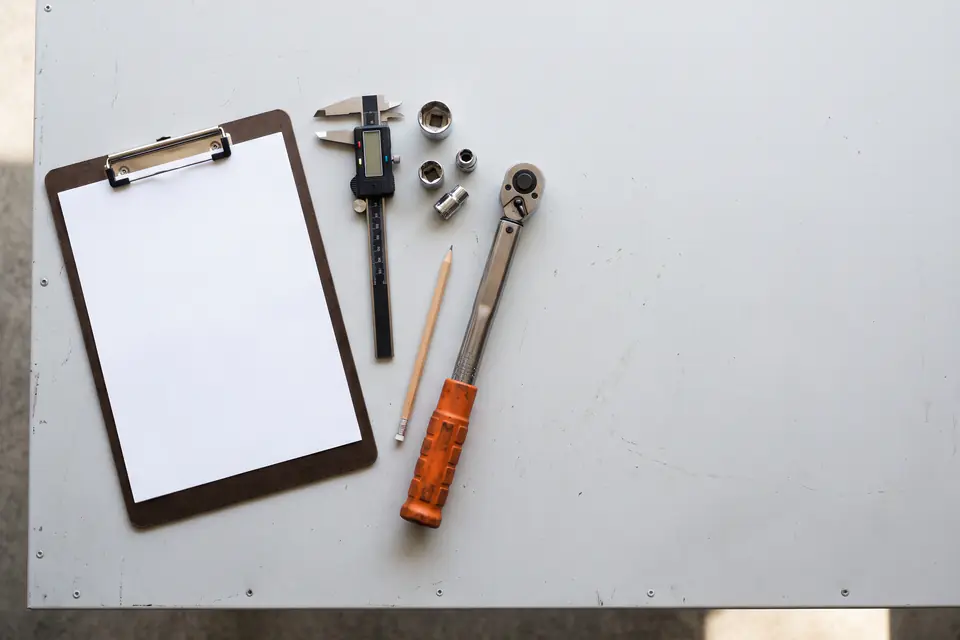 A clipboard with a blank sheet, a digital caliper, sockets, a pencil and a ratchet on a gray workbench, seen from above.